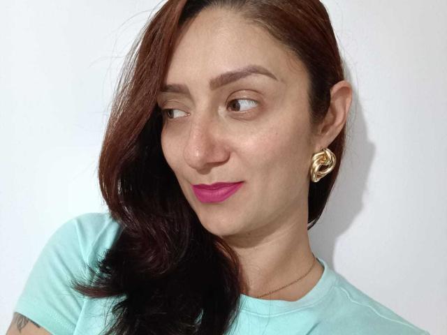 ValenRoseM has brown hair, pink lipstick, and gold hoop earrings, wearing a light green shirt, looking sideways with a slight smile. She has a small tattoo on her arm.