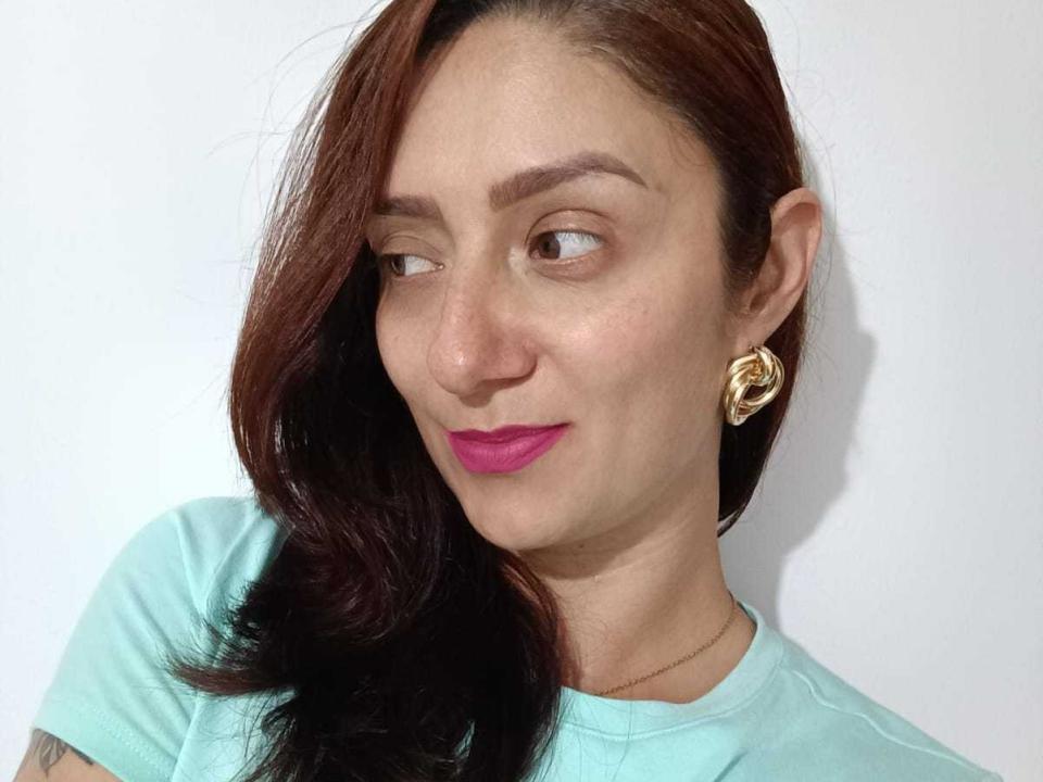 ValenRoseM has brown hair, pink lipstick, and gold hoop earrings, wearing a light green shirt, looking sideways with a slight smile. She has a small tattoo on her arm.