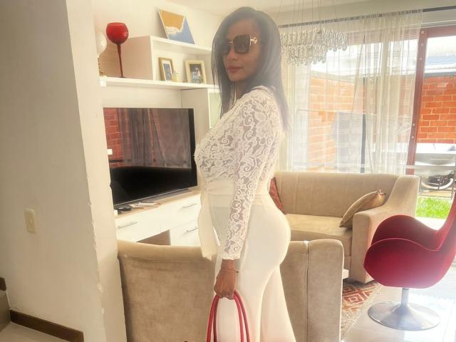 Anahismaar with long black hair, wearing white lace long-sleeve top and tight white pants, holding red straps, standing in modern living room, wearing sunglasses, showcasing her ass.