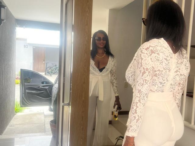 Anahismaar with dark skin, wearing a white lace long-sleeve top and pants, standing in a doorway, holding a bottle, looking at another Anahismaar with similar attire and black hair.