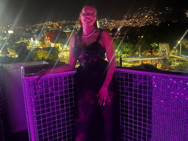 Anahismaar with purple-lit tits, wearing a black lace dress, pearl necklace, and red earrings, leans on a tiled balcony, city lights in the background, night, smiling.