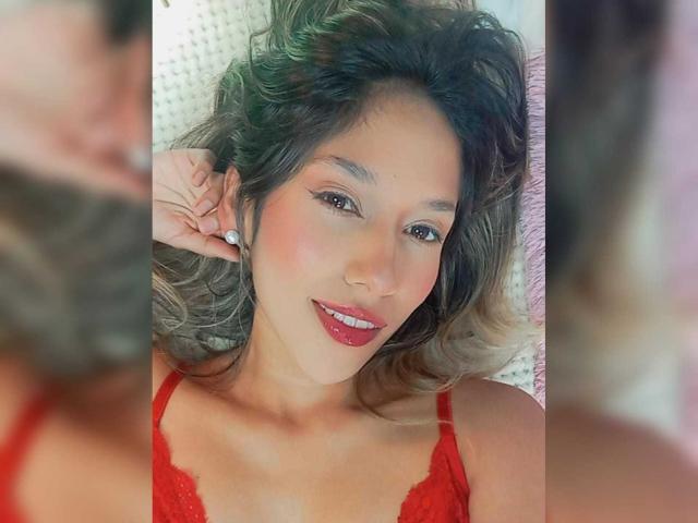 BrianaHarpers tem cabelos castanhos ondulados, batom vermelho e está usando um sutiã de renda vermelho, sorrindo com a mão perto da orelha. O reflexo ao fundo está borrado..