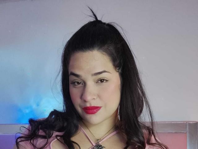 AntonelaPark with dark hair in a high pony, red lipstick, pink top, and necklace, looking at camera, soft smile, light skin, slightly wavy hair, blue light in background.