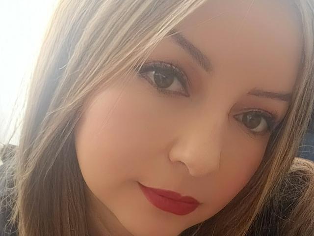 NatalyHami has brown eyes, light skin, straight brown hair, and red lipstick. She's looking at the camera with a slight smile. Her nose is small and she's cute.