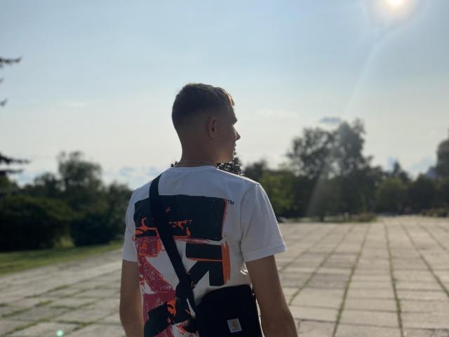 PeterFortune, vu de dos, portant un t-shirt blanc avec des motifs colorés, un sac bandoulière noir, debout sur un chemin carrelé ensoleillé, regardant à droite, le soleil brille d'en haut..