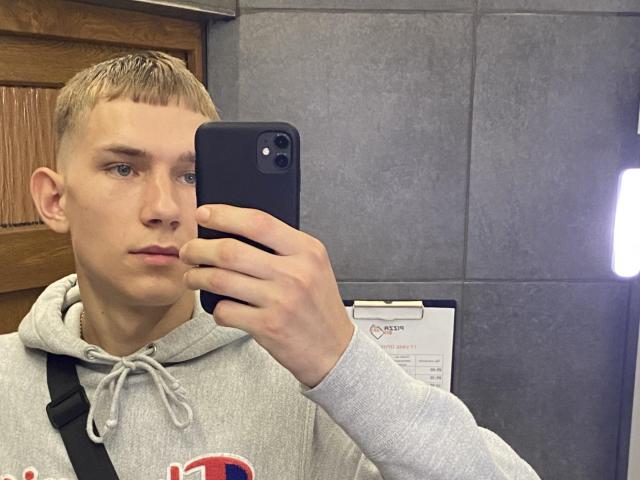 PeterFortune, un niño rubio con cabello corto, sosteniendo un teléfono negro en un selfie frente al espejo, vistiendo una sudadera gris, expresión neutra, pared de baldosas grises, puerta de madera, correa negra, luz proveniente de la derecha..