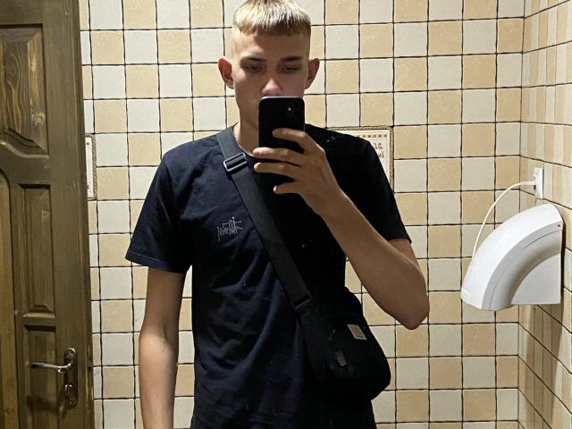 PeterFortune, cheveux blonds et courts, t-shirt noir, sac bandoulière noir, tenant un téléphone, autoportrait avec un miroir, porte en bois, mur carrelé, distributeur de serviettes en papier blanc sur la droite..