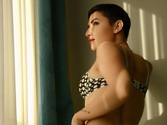 AmyOffice with short black hair, polka dot bra, and tattoo on right wrist, standing by window, sunlight casting shadows, looking over shoulder, red lipstick, confident, sexy.