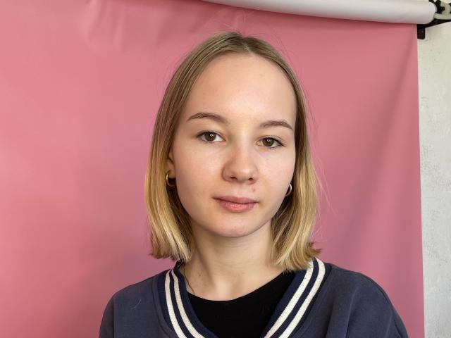 Tracy Marshall heeft blond haar dat tot op haar schouders reikt, een lichte huid, een kleine neus, een subtiele glimlach, bruine ogen, draagt een marineblauwe blouse met witte strepen, een zwarte top, kleine oorbellen en heeft een roze achtergrond..