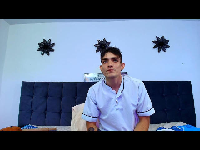Keytwoo is a young dude with short black hair, wearing a white polo shirt, sitting on a bed with a blue tufted headboard, and black flower wall decorations.