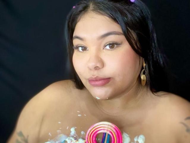 CarolineHardman with black hair, brown eyes, and tan skin, has pink and white candy swirls on her chest and face, holding a pink and white candy swirl in her hands.