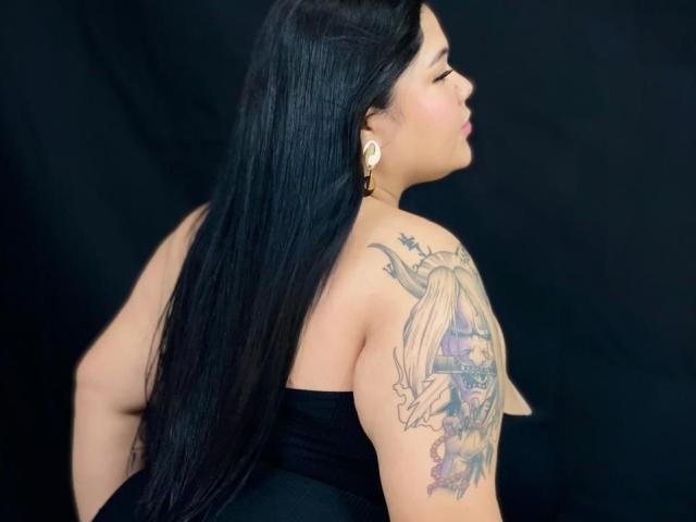 CarolineHardman with long black hair, facing side, showing tattoo on upper arm, wearing black strapless dress, gold earrings, black background, tattoo features horned woman and text.