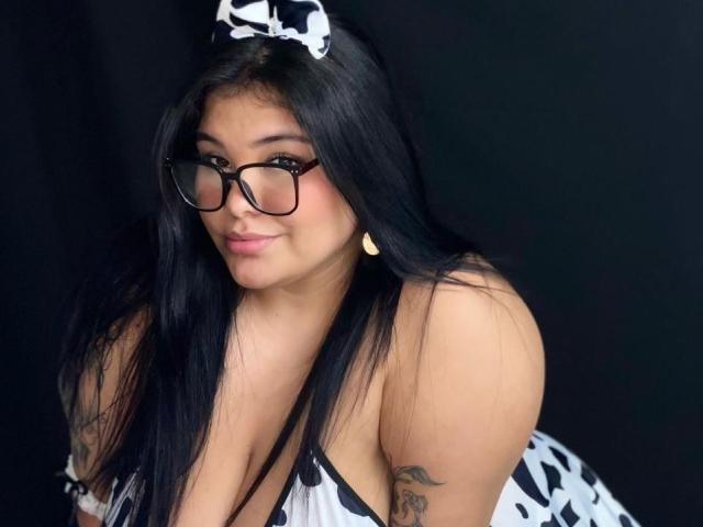 CarolineHardman with long black hair, glasses, cow-patterned outfit, and cow ears, has a cute smile, small gold earrings, tattoo on right arm, and large, curvy, bare breasts.