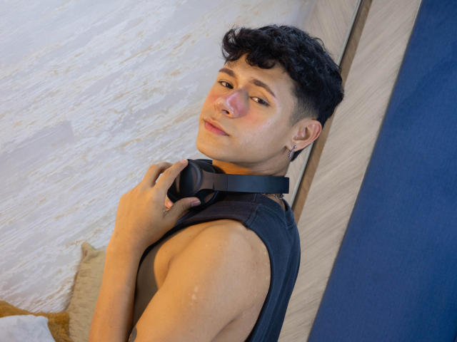 DanteEvans with curly black hair, wearing a sleeveless black shirt, holding headphones around his neck, looking over shoulder, pale skin, small nose, slightly flushed, ear piercing visible.