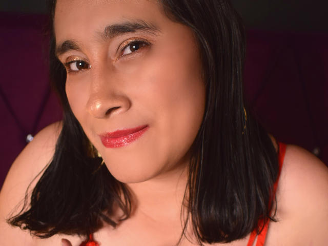EvaChain tem uma pele marrom brilhante, cabelo preto e liso, olhos castanhos e batom vermelho. Ela está vestindo uma blusa vermelha e brincos dourados, com um sorriso discreto. Seus seios são pequenos..