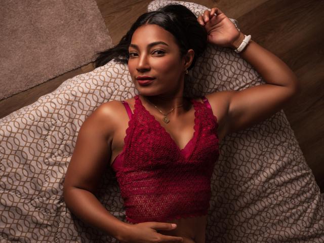EmmaColeman69 laying on a bed, wearing a red lace crop top, showing her tits, dark skin, black hair, looking at the camera, hand on her stomach, wooden floor.