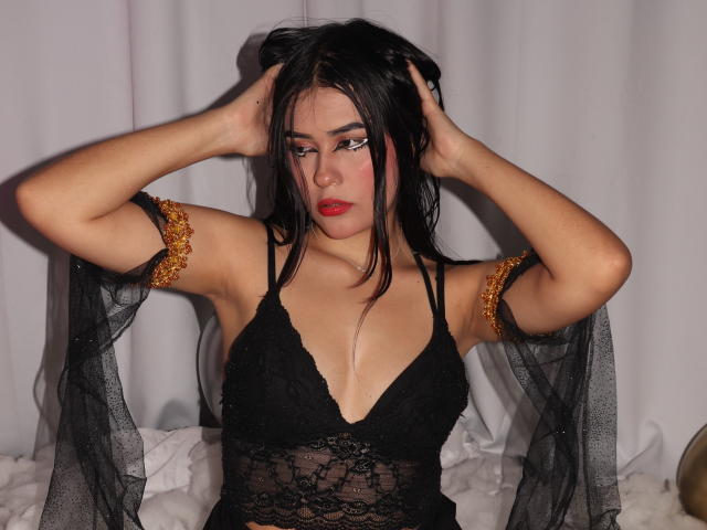 JulietaGarcia with black lace lingerie, arms raised, hands in hair, red lipstick, black hair, gold arm bands, sheer black fabric, white background, looking down, medium tits.