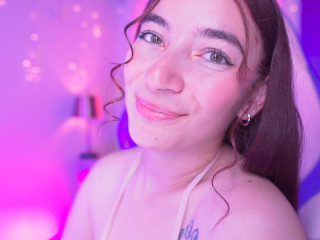 IssaMonterrey with curly brown hair, pinkish lips, green eyes, and a subtle smile, wearing a white halter top, showing a small tattoo on her right shoulder.