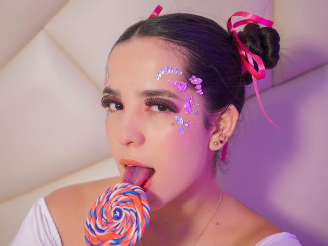 AlanaReyes with makeup glitter on her cheek, tongue out, licking a colorful swirl candy, pink ribbons in her black hair, white shirt, and earrings, in a soft-lit photo.