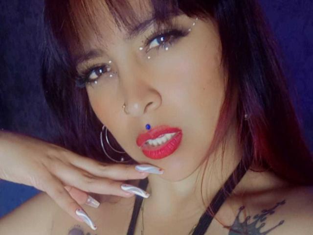 Sarah69Hot has dark brown hair, red lipstick, long silver nails, a nose ring, hoop earrings, and a small blue dot above her lip. She's looking seductively at the camera.