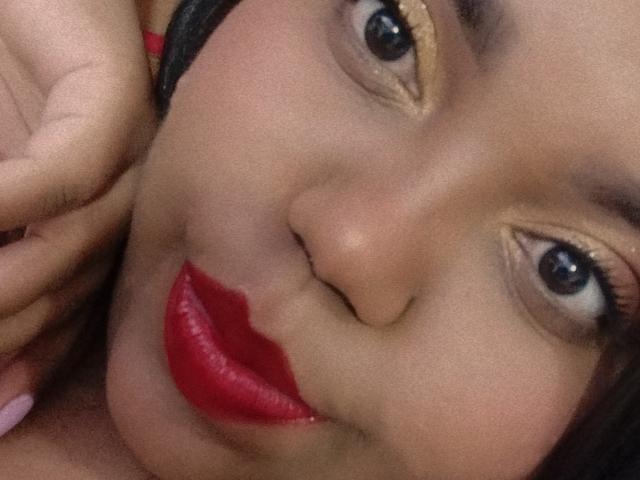 SarahSmity's face with glossy red lips, gold eyeliner, and black hair, touching her cheek with a hand, showing pink nails, and looking at the camera with a slight pout.