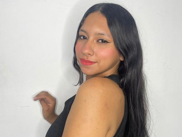 AshleyRosel has long black hair, wearing a black sleeveless top, looking over her shoulder with a slight smile, red lipstick, and black eyeliner, against a white wall.