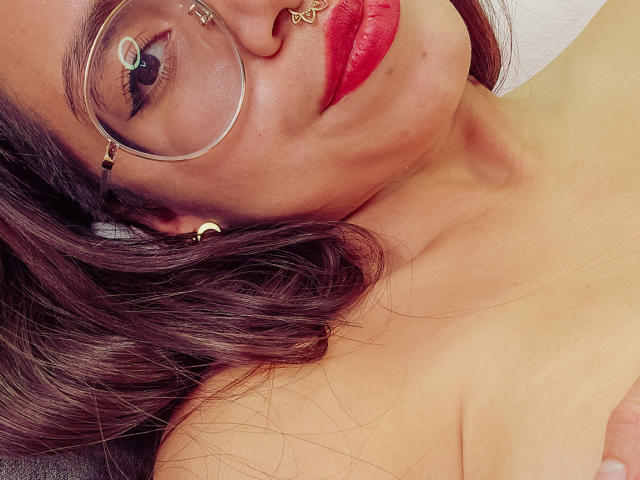 NaturalMariaHun's face with red lipstick, glasses, and gold earrings, lying down, showing a bit of her bare shoulders and brown wavy hair, looking up with a slight smile.