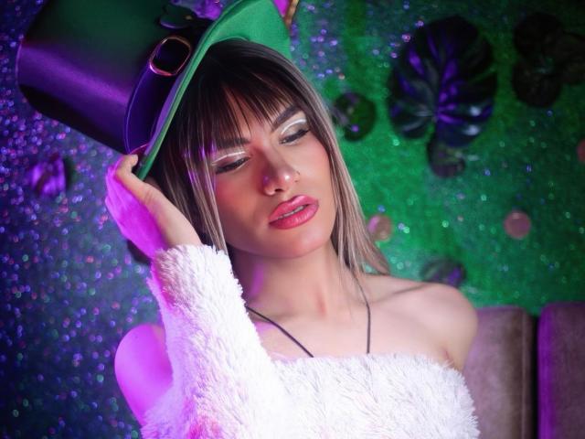 ZamaraKleii with pink lipstick, green hat, white fuzzy top, holding hat with left hand, purple-green lighting, sparkling background, straight brown hair with bangs, looking slightly down.