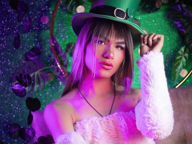 ZamaraKleii wearing a green top hat with a black band, pink fluffy off-shoulder shirt, purple and green lighting, holding hat, long brown hair, pink lips, sexy look.