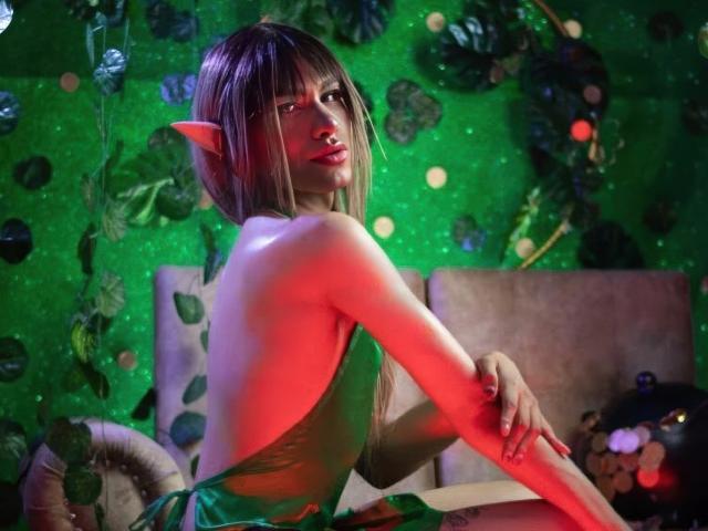 ZamaraKleii with pointy elf ears, green dress, backless, long blonde hair, red light, green background, leaves, and glowing orbs, sitting, looking over shoulder, sexy, attractive, cute.