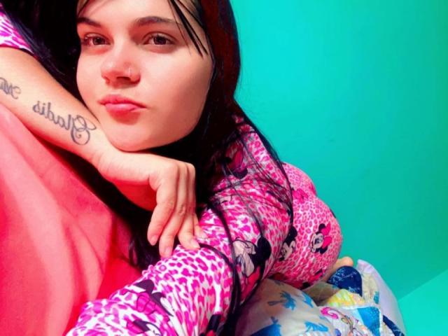 GalaLaverne with dark hair, pink leopard print shirt, tattoo on right wrist, and pink bedsheet, leaning on her hand, looking at camera, against teal wall.