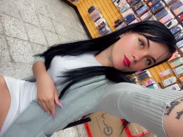 GalaLaverne with long black hair, wearing a white top and gray cardigan, showing her pink-nail-painted hand, white panties, and a tattoo on her right arm, taking a selfie.