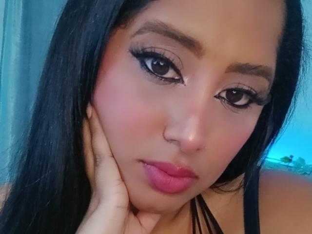 MariaCatalina's face, with pink lips, black eyeliner, and long black hair, resting her hand on her cheek, showing a cute, sexy, and slightly flirty expression.