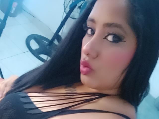 MariaCatalina with black hair, pink lips, and heavy eyeliner, wearing a strappy black top, pouting her full, round boobs in a selfie.