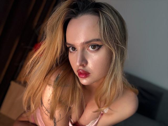 TurboHottie with shiny skin, blonde hair, and red lips, looking up with black eyeliner. She's wearing a pink top, showing her tits, and has a sexy, glistening face.