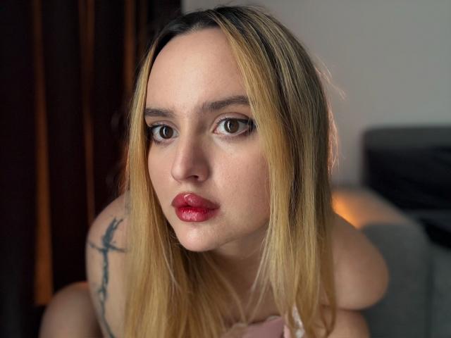 Eine attraktive Frau mit blonden Haaren, roten Lippen und Tattoos an der Schulter, die direkt in die Kamera mit braunen Augen schaut, trägt ein pinkes Oberteil und befindet sich in einem sanft beleuchteten Raum..
