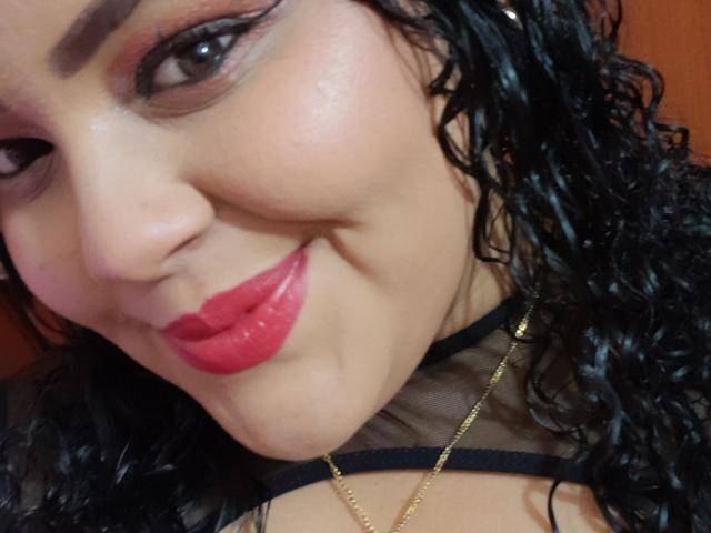 MelannieCarter's face, with curly black hair, red lipstick, and a gold necklace, smiling slightly. Her brown eyes have winged eyeliner, and she wears a sheer black top.