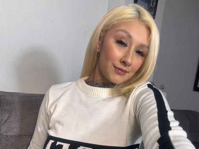 RachellGrey with blonde hair, wearing a white sweater with black stripes, has tattoos on her neck, and is smiling slightly at the camera. Her tits are small and she's cute.
