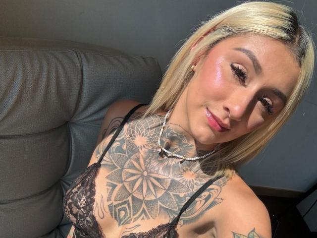 RachellGrey, con cabello rubio, pecho tatuado y un sujetador de encaje negro, sonriendo a la cámara, iluminada por el sol, llevando un collar de perlas, luciendo sexy..