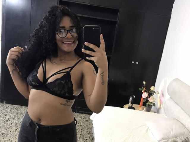 ThiainaRose met krullend zwart haar, een bril, een zwarte kanten bh, kleine borsten, een tatoeage op haar rechterarm, die een telefoon vasthoudt, lachend, gekleed in zwarte broek, staand in een slaapkamer, spiegelzelfportret..
