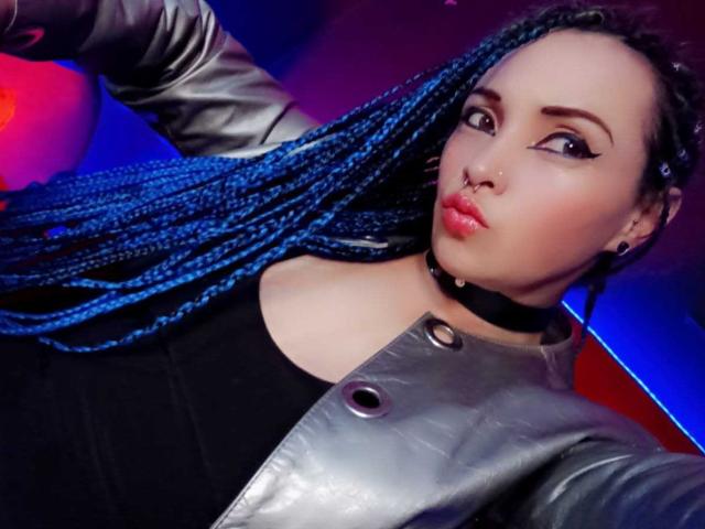 Dalila Musa, con trenzas azules, labios carnosos, collar negro, chaqueta de cuero y los pezones al descubierto, mirando hacia un lado. Lleva maquillaje con delineador de ojos llamativo..