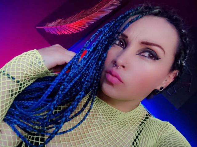 Dalila Musa mit blauen Zöpfen, Netzoberteil, Nasenring und schwarzen Ohrringen, die die Lippen zusammenpresst und einen ernsten Gesichtsausdruck zeigt, während sie ihr Haar hält. Ihre Brüste sind durch das Netz sichtbar..