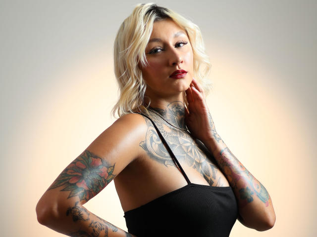 RachellGrey, con capelli biondi, tatuaggi sulle braccia e sul petto, indossa una maglietta nera, con la mano vicino al collo, guarda la fotocamera, rossetto rosso, illuminazione soffusa, leggero sorriso, posa sicura..