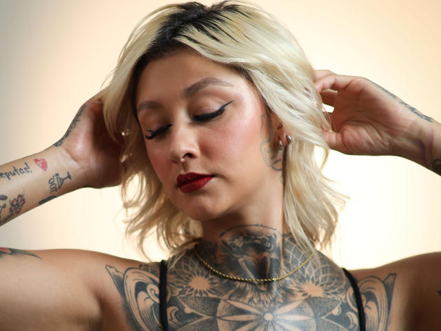 RachellGrey met blond haar, tatoeages op haar nek en armen, rode lippenstift, zwarte eyeliner, handen in haar haar, een gouden ketting, die haar tatoeages op haar borsten en billen laat zien in een portret..