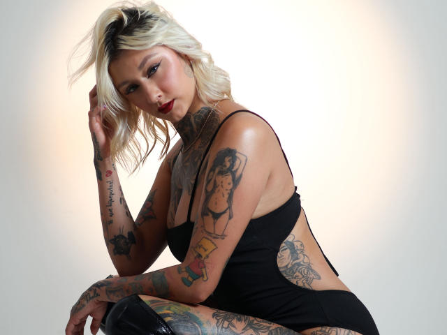 RachellGrey met platinablond haar, tatoeages op armen en borst, draagt een zwart tanktopje, zit, kijkt naar de camera, heeft haar hand in haar haar, rode lipstick, zwarte kniehoge laarzen..