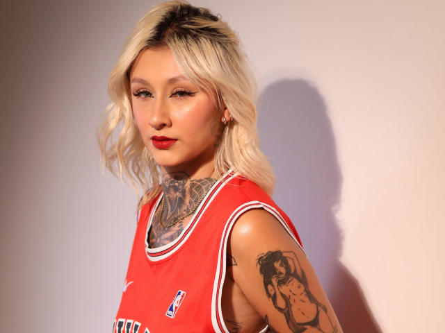 RachellGrey, con cabello rubio, lápiz labial rojo, tatuajes en el cuello y el brazo, vestida con una camiseta roja de baloncesto, mirando a la cámara con una expresión seria. La luz del sol proyecta una sombra..