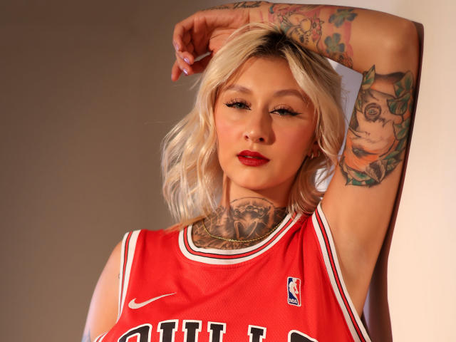 RachellGrey met blond haar, rode lippenstift, tatoeages op arm en nek, gekleed in een rood basketbalshirt, arm omhoog, kijkend naar de camera, zachte belichting, aantrekkelijk..
