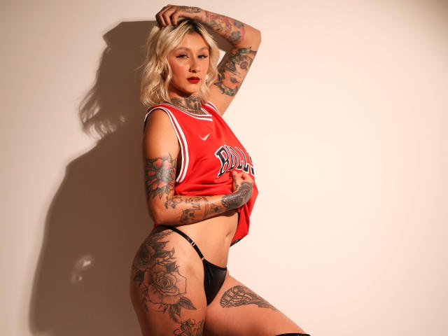 RachellGrey, con cabello rubio, tatuajes, una camiseta deportiva roja de Nike, tanga negra y lápiz labial rojo, de pie con el brazo levantado, mostrando su trasero curvilíneo y sus tatuajes..