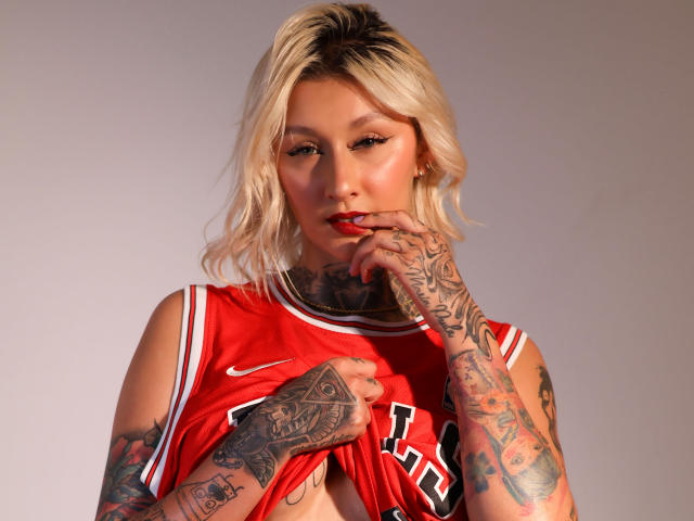 RachellGrey, con cabello rubio, tatuajes en manos y brazos, vestida con una camiseta roja de baloncesto, levantándola para mostrar sus pequeños pezones perforados, mirando a la cámara con un dedo en el labio..