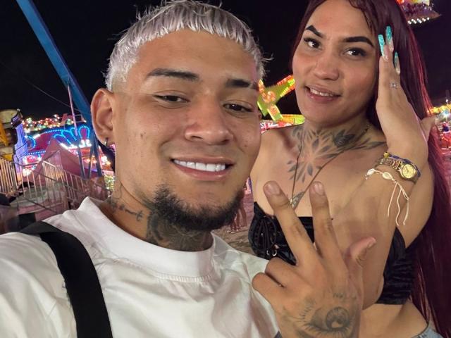 MegaSexyHot with silver hair, tattoos, and white tee, holding MegaSexyHot with long red hair, big tits, black top, and blue nails, flashing a peace sign at night.